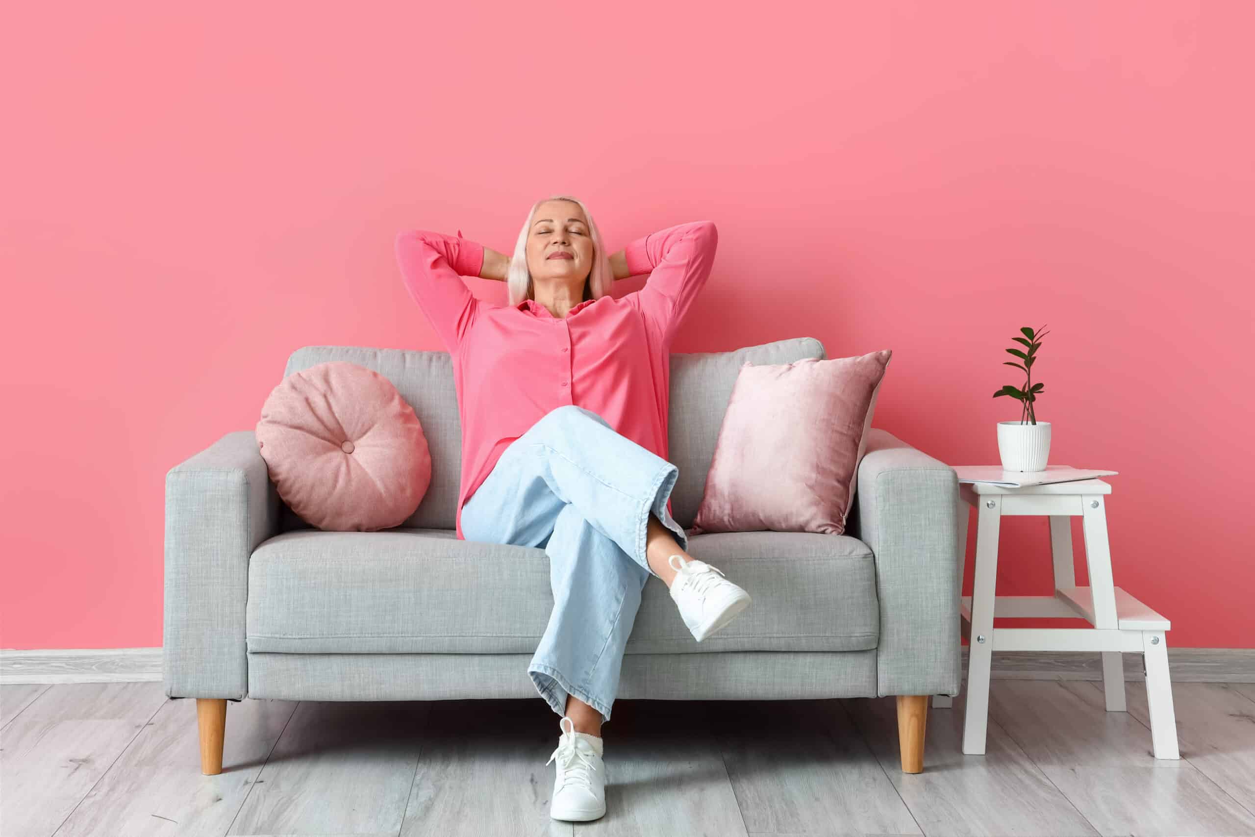 Mature woman relaxing on a gray couch in front of a pink wall.